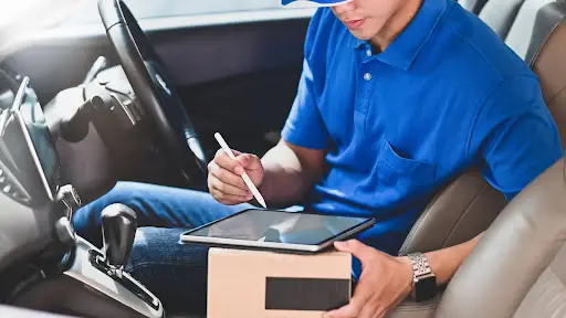 A delivery driver in a blue shirt and cap sits in a car, holding a small package and using a stylus on a tablet, likely confirming a delivery or updating order status via PerGo transportation management software.