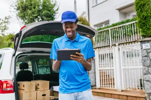 A smiling delivery person in a blue shirt and cap uses a tablet powered by PerGo transportation dispatch software while standing beside an open car trunk filled with boxes in a residential area.