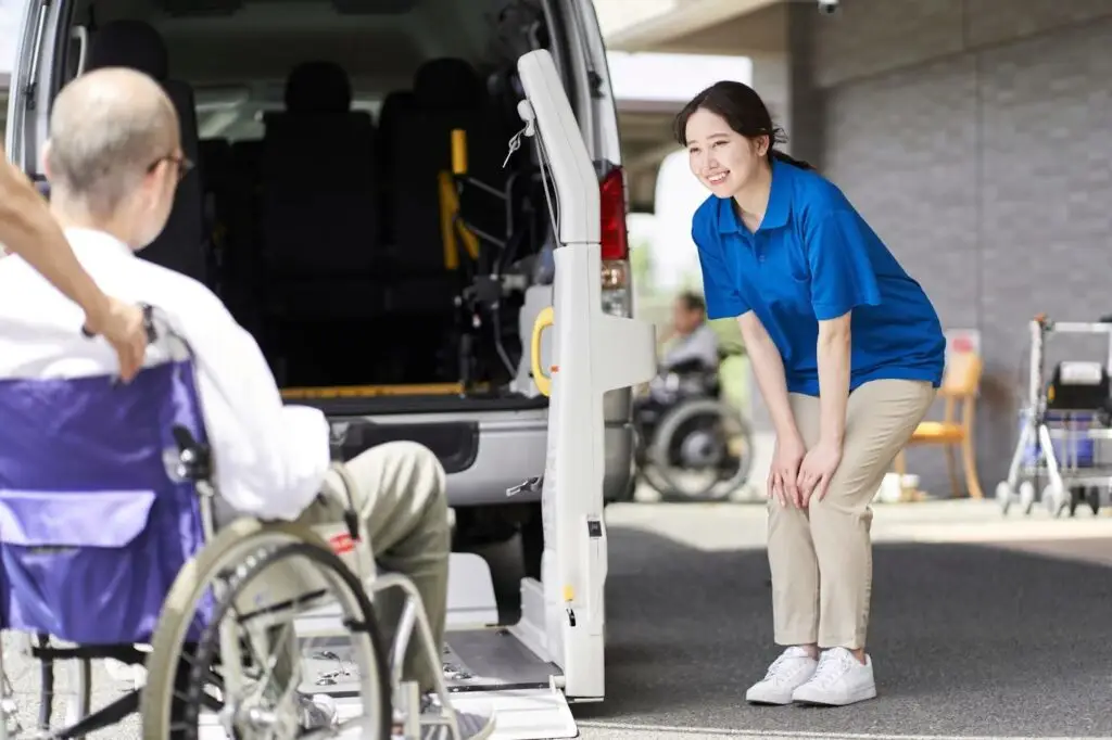 A caregiver in a blue shirt smiles while assisting a person in a wheelchair near a van with a wheelchair lift, showcasing how PerGo NEMT Software streamlines accessible transportation on a sunny day.