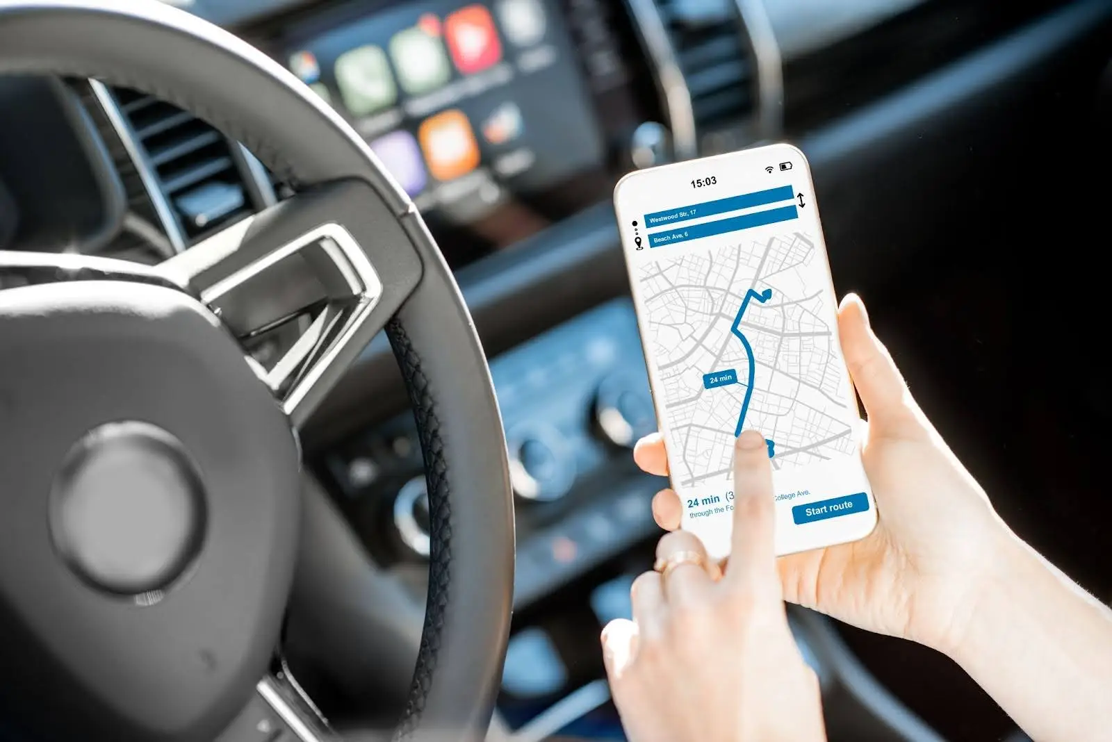 A person in a car uses a smartphone with a navigation app powered by PerGo transportation dispatch software. The car&rsquo;s steering wheel and dashboard are visible in the background.