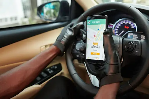 A person wearing black gloves holds a smartphone showing a ride-sharing app with a “New Request” notification powered by transportation dispatch software while driving a car; the dashboard and steering wheel are visible.