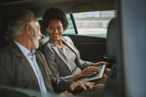 Two business professionals, a man and a woman, sit in the back seat of a car. The woman uses her laptop with PerGo transportation management software, smiling at her colleague as they discuss seamless collaboration and efficient travel planning.