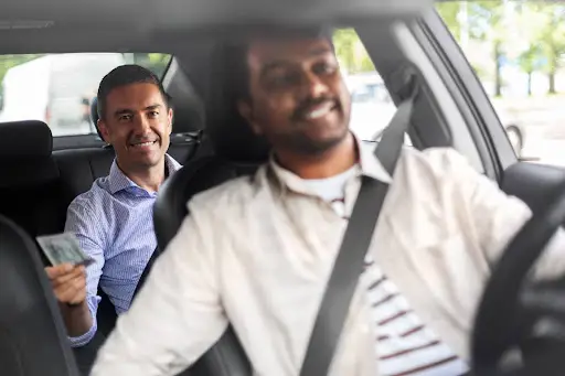 A smiling passenger in the back seat hands cash to a cheerful driver in the front of a car during a daytime ride, showcasing the convenience made possible by efficient transportation dispatch software.