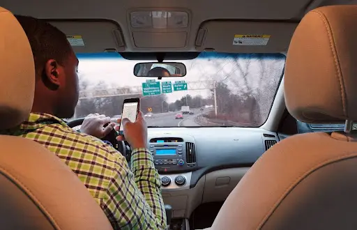A man driving a car is looking at his phone while holding the steering wheel. The photo, taken from behind the front seats, highlights how NEMT Software can enhance safety and efficiency in transportation dispatch software solutions.