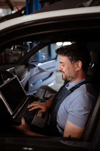 A man sitting in the driver's seat of a car uses a laptop, appearing to run diagnostics or perform maintenance with PerGo NEMT Software inside a garage or workshop.