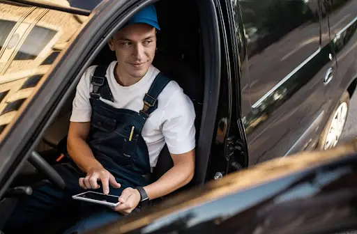 A person wearing a white shirt, blue overalls, and a blue cap sits in the driver's seat of a car, smiling and using a tablet with PerGo transportation dispatch software, as a city street is reflected in the car&rsquo;s window.
