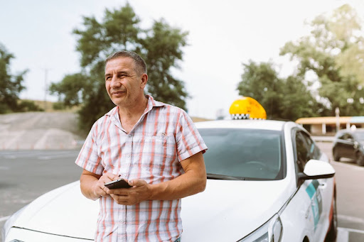 A man in a plaid shirt stands in front of a white taxi, holding a smartphone. Trees and a road are visible in the background on a sunny day, highlighting the convenience of NEMT Software like PerGo for seamless transportation management.
