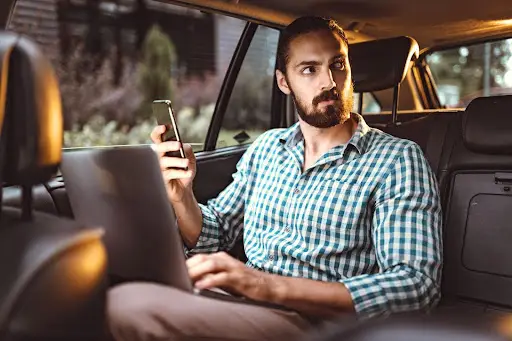 A man with a beard, wearing a checkered shirt, sits in the back seat of a car with his laptop on his lap and phone in hand, looking out the window with a serious expression as he manages tasks using transportation dispatch software.