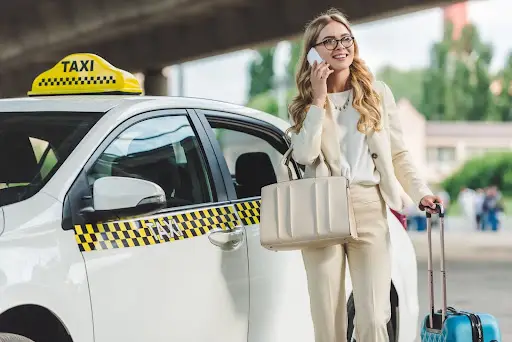 A smiling woman in business attire stands beside a white taxi, talking on her phone and holding a suitcase and a large handbag&mdash;perhaps coordinating her ride with PerGo, advanced transportation management software. Trees and a bridge are visible in the background.