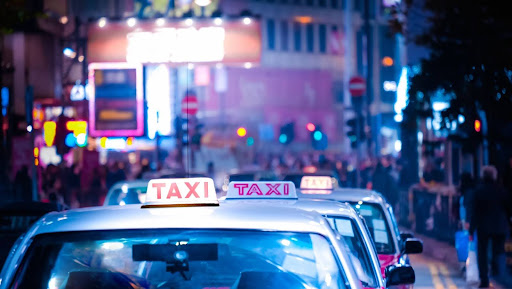 Taxis with illuminated signs wait in line on a busy city street at night, surrounded by vibrant neon lights and blurred crowds&mdash;seamlessly coordinated thanks to advanced transportation dispatch software like PerGo.