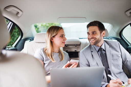 Two business professionals, a woman and a man in formal attire, sit in the backseat of a car, smiling and talking. The woman holds a phone displaying PerGo Transportation Management Software while the man works on his laptop with a pen.