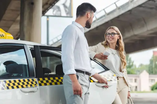 A man holds open a taxi door for a smiling woman standing nearby. Under an overpass, they chat as the woman, in business attire, checks her smartphone&mdash;possibly coordinating her ride with transportation dispatch software.