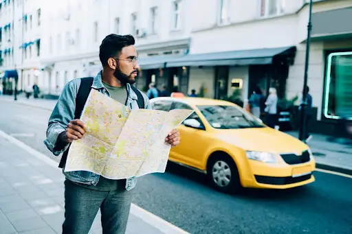 A man with glasses and a backpack stands on a city sidewalk, holding an open map and looking to the side, while a yellow taxi&mdash;possibly managed by transportation dispatch software&mdash;passes by on the street behind him.