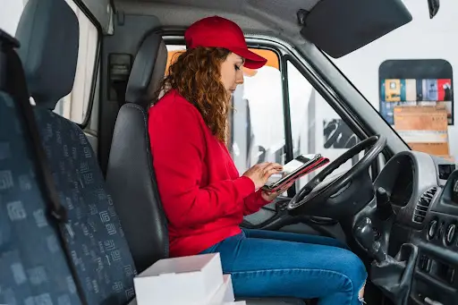 A delivery driver in a red cap and sweatshirt sits in a van's driver's seat, using transportation dispatch software on her tablet. Several packages are visible on the passenger seat next to her.