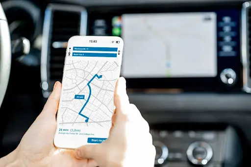 A person holds a smartphone displaying a navigation map with a route, inside a car. The dashboard and built-in GPS screen are visible, highlighting how PerGo NEMT software streamlines transportation dispatch for efficient journeys.