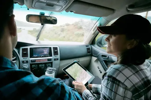 A woman in a plaid shirt and cap sits in the passenger seat of a car, smiling and holding a tablet with a map powered by transportation dispatch software. A man drives, and the dashboard displays another map.