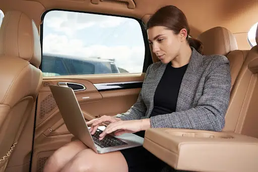A woman in business attire works on a laptop while sitting in the back seat of a car with tan leather interior, appearing focused as she manages tasks with PerGo transportation dispatch software.