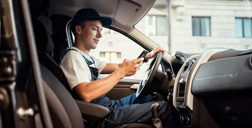 A man wearing a cap and work overalls sits in the driver&rsquo;s seat of a van, holding the steering wheel with one hand and checking transportation dispatch software on his smartphone with the other.