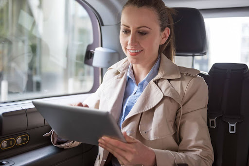 A woman wearing a beige trench coat sits in the backseat of a car, smiling while using a tablet with transportation dispatch software, as natural light filters through the window.