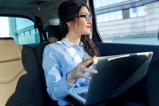 A woman with glasses and a blue shirt sits in the backseat of a car, smiling and looking out the window while holding an open laptop on her lap, possibly using PerGo transportation management software.