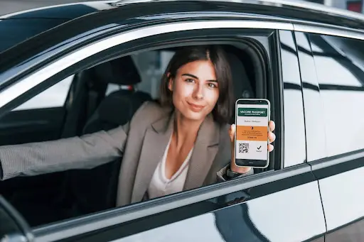 A woman sitting in a car holds out her smartphone, displaying a digital vaccine passport with a QR code. She smiles slightly, ready to show her proof of vaccination&mdash;making travel and Transportation management Software like PerGo more efficient than ever.
