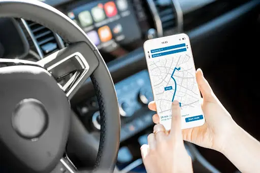 A person sitting in a car uses a smartphone with PerGo NEMT Software displaying a route on a map, while holding the steering wheel with the other hand. The car dashboard and controls are visible in the background.