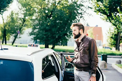 A man with a beard stands next to an open taxi door, holding a smartphone and wearing earphones, possibly using transportation dispatch software. He is outdoors on a sunny day with trees and buildings in the background.