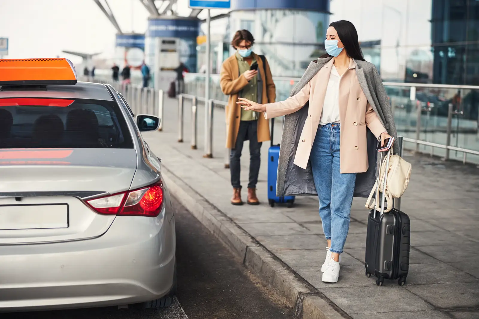 A woman wearing a mask and holding luggage signals for a taxi outside an airport, while another masked person with a suitcase stands behind her checking their phone&mdash;modern travel scenes made smoother with transportation dispatch software.