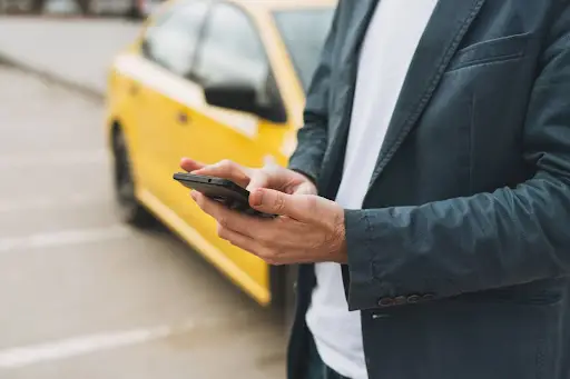 A person in a suit uses a smartphone with PerGo transportation dispatch software while standing next to a parked yellow car in an outdoor parking lot. The focus is on the person's hands and phone.