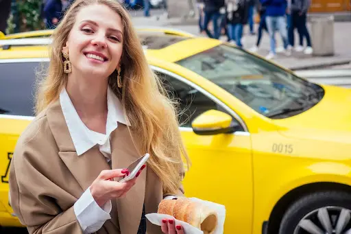 A smiling woman with long blonde hair stands in front of a yellow taxi, holding a phone&mdash;perhaps using transportation dispatch software&mdash;in one hand and a pastry in the other. She is wearing a tan coat over a white shirt, with people and city buildings visible in the background.