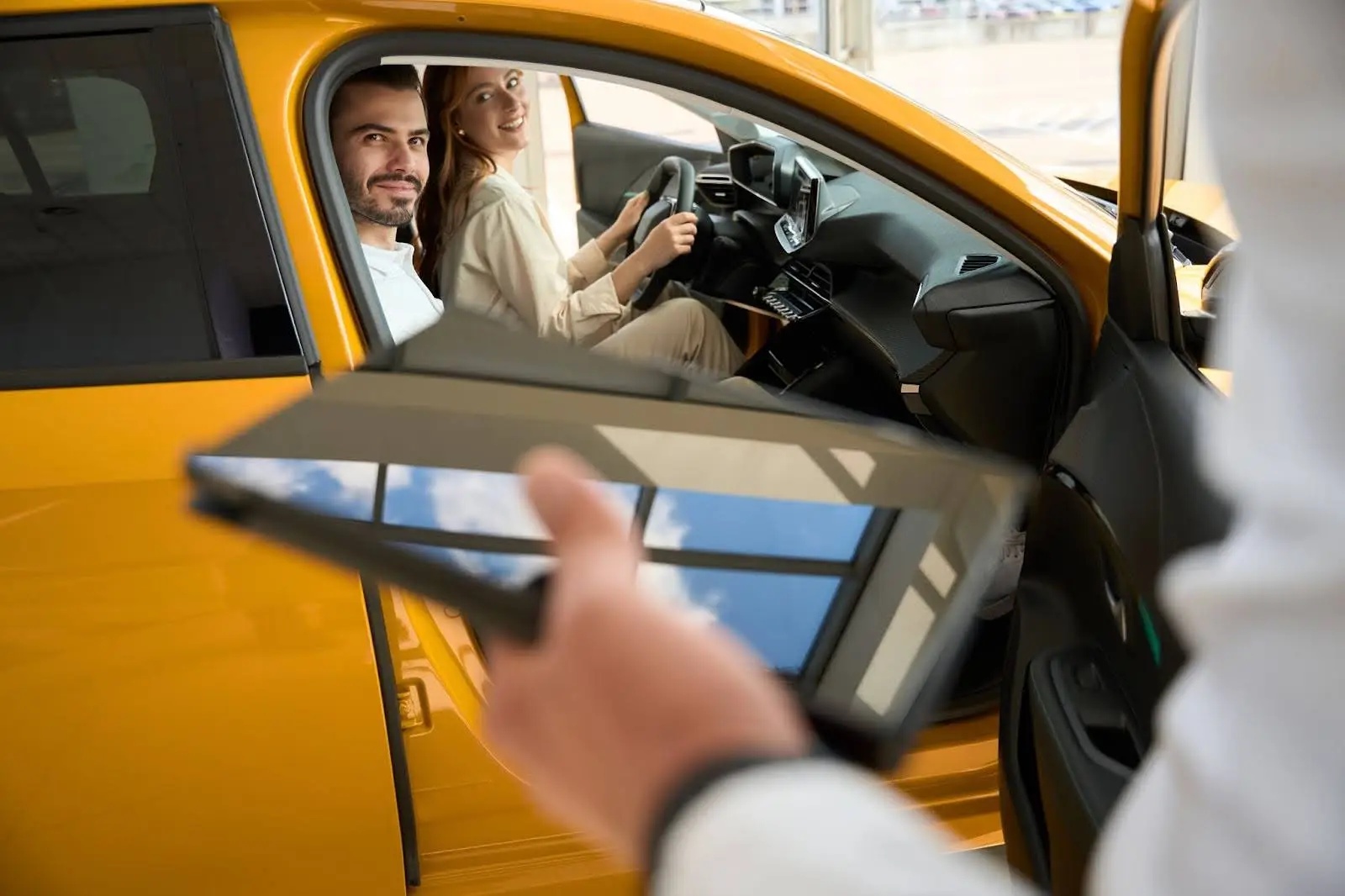 A man and woman sit in the front seats of a yellow car, smiling, while a person holding a tablet&mdash;possibly using PerGo transportation dispatch software&mdash;stands outside the open driver&rsquo;s door, likely at a car dealership.