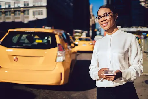 A woman wearing glasses and a white blouse stands on a city street, smiling and holding a smartphone. Yellow taxis in the background hint at an urban setting, possibly managed efficiently with transportation dispatch software.