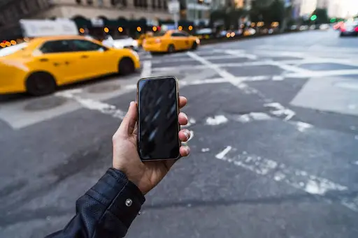 A person holding a smartphone with a blank screen on a city street, yellow taxis driving by, and buildings lining the road&mdash;perfect for showcasing PerGo&rsquo;s transportation dispatch software in action.
