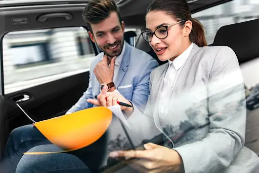 A woman in business attire with glasses is showing a presentation on NEMT software to a man beside her while sitting in the back seat of a car. Both are engaged and appear to be discussing work-related features of the platform.