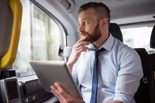 A man with a beard sits in a car, holding a tablet running NEMT Software, and looks thoughtfully out the window. He is wearing a light blue shirt and a loose tie, with one hand touching his chin.