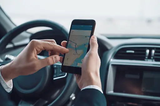 A person sitting in a car uses a smartphone displaying a map app powered by PerGo Transportation Management Software, with one hand holding the phone and the other hand pointing at the screen. The steering wheel is visible in the background.