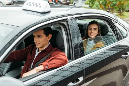 A woman sits in the back seat of a black taxi, smiling, while the male driver focuses on the road&mdash;efficiently navigating with PerGo transportation dispatch software. Trees and parked cars pass by outside.