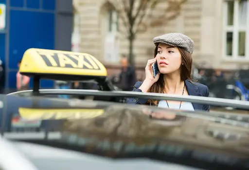 A woman wearing a flat cap talks on her phone while standing next to a taxi on a city street, possibly coordinating a ride with transportation dispatch software. The yellow taxi sign is visible on the car roof in the foreground.
