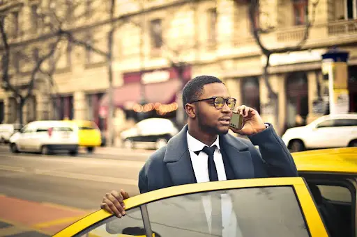 A man in a suit and glasses stands by an open yellow car door, talking on his phone&mdash;perhaps coordinating with PerGo transportation dispatch software&mdash;with a city street and blurred buildings in the background.