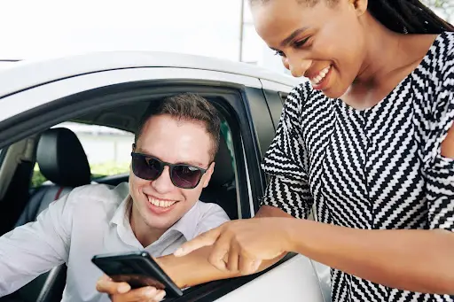 A smiling man wearing sunglasses sits in a car while a woman standing outside shows him something on her smartphone&mdash;both appear happy and engaged, possibly exploring transportation dispatch software together.