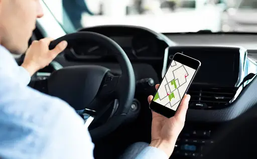 A person sitting in a car is holding a smartphone with a navigation map powered by PerGo transportation dispatch software, while their other hand is on the steering wheel. The view is from inside the car.