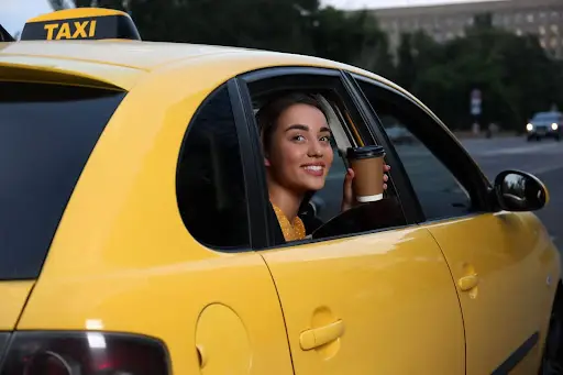 A woman smiles while holding a coffee cup, sitting in the backseat of a yellow taxi with the window down&mdash;enjoying a smooth ride managed by advanced transportation dispatch software. Trees and buildings are visible outside.
