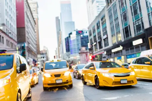 Several yellow taxis drive down a busy city street surrounded by tall buildings, showcasing the efficiency of transportation dispatch software amid blurred vehicles and pedestrians in a bustling urban environment.