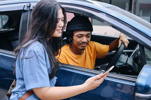 A woman stands outside a blue car, smiling and showing her phone&mdash;displaying advanced transportation dispatch software&mdash;to a man in an orange shirt and black beanie who is sitting in the driver&rsquo;s seat; both appear happy and engaged.