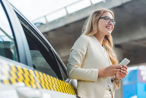 A smiling woman in business attire stands by a taxi, holding a smartphone, showcasing the ease of transportation dispatch software under a blue sky and overpass.