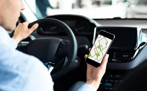A person driving a car holds a smartphone displaying a navigation route on PerGo transportation dispatch software, glancing at the screen while keeping one hand on the steering wheel.