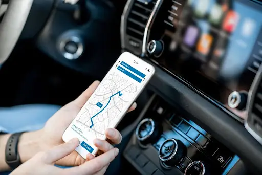 A person holding a smartphone with PerGo transportation dispatch software open, displaying a map and route, while sitting in the driver&rsquo;s seat of a car with the dashboard and touchscreen visible.
