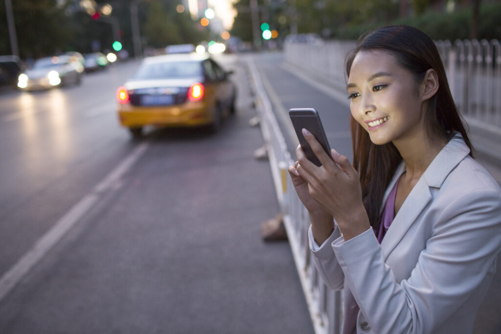A woman in a light blazer stands on a city sidewalk at dusk, smiling while using her smartphone and the PerGo transportation dispatch software. A taxi is parked nearby, with cars and streetlights illuminating the background.