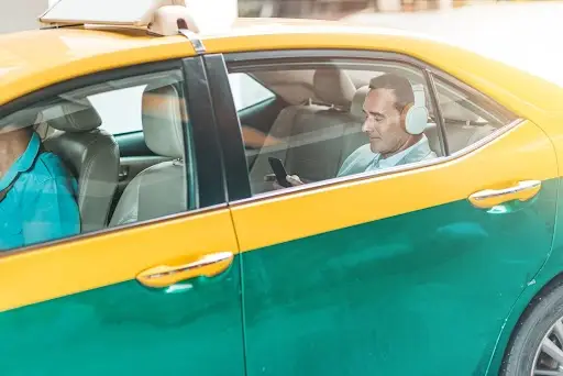 A man wearing headphones sits in the back seat of a yellow and green taxi, looking at his phone, while another person drives the car.
