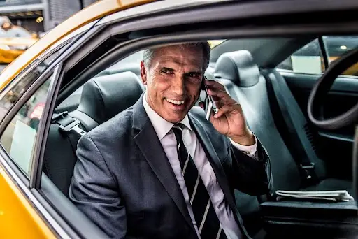 A man in a suit sits in the back seat of a car, smiling while talking on a smartphone. A newspaper is beside him, and the car door is open, showing part of a yellow vehicle exterior.